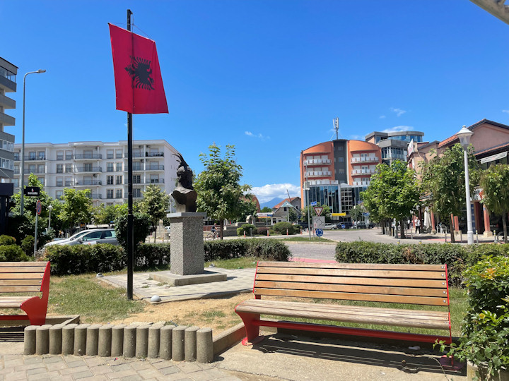 A town square with park benches, an Albanian flag, and a monument.