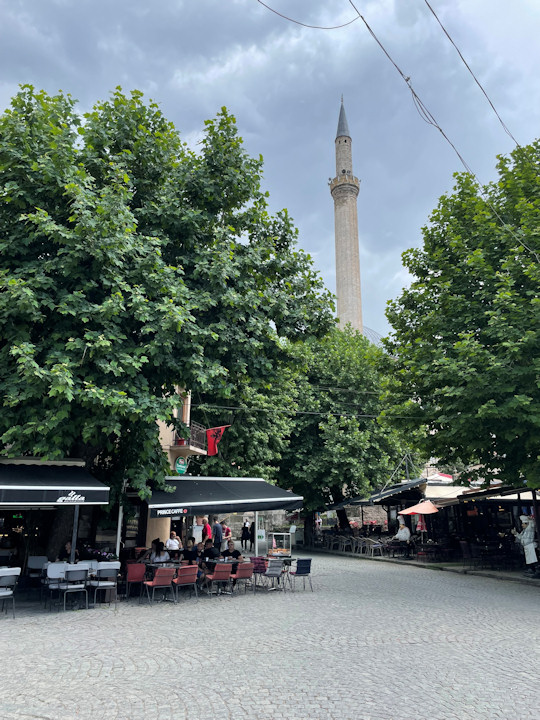 A cobblestone town square with street cafes, trees, and a minaret rising to the sky.